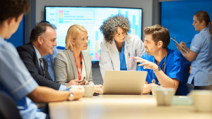A diverse group of medical professionals and business people gather around a laptop, discussing earnestly. Background features data charts. Collaborative mood.