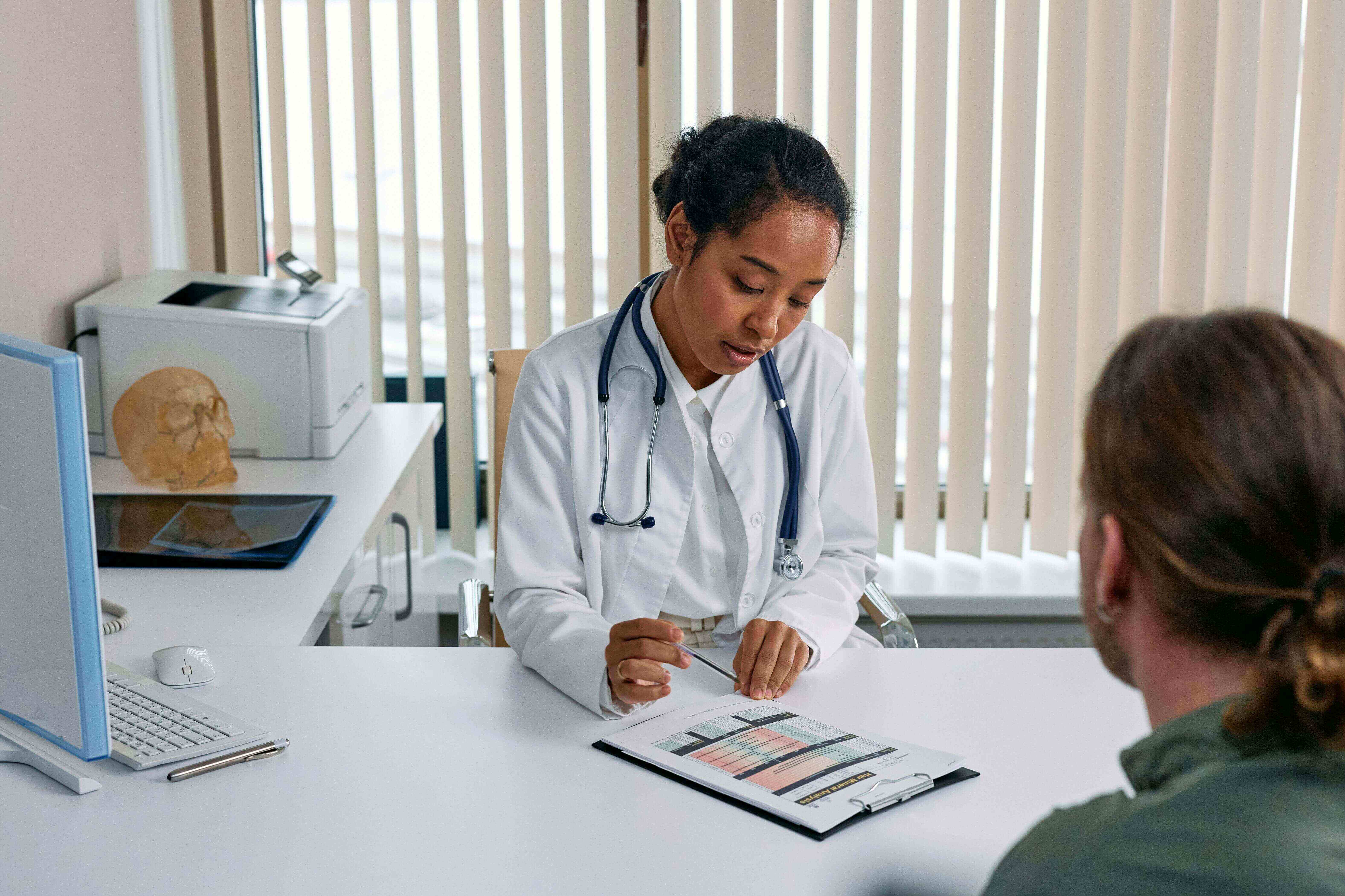 A doctor with a stethoscope reviews a clipboard with a patient in an office. A computer and model skull are on the desk, blinds in the background.