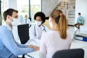 A doctor converses with a masked couple in a bright medical office. The couple holds hands, expressing support. Background shows others waiting, creating a calm, focused atmosphere.
