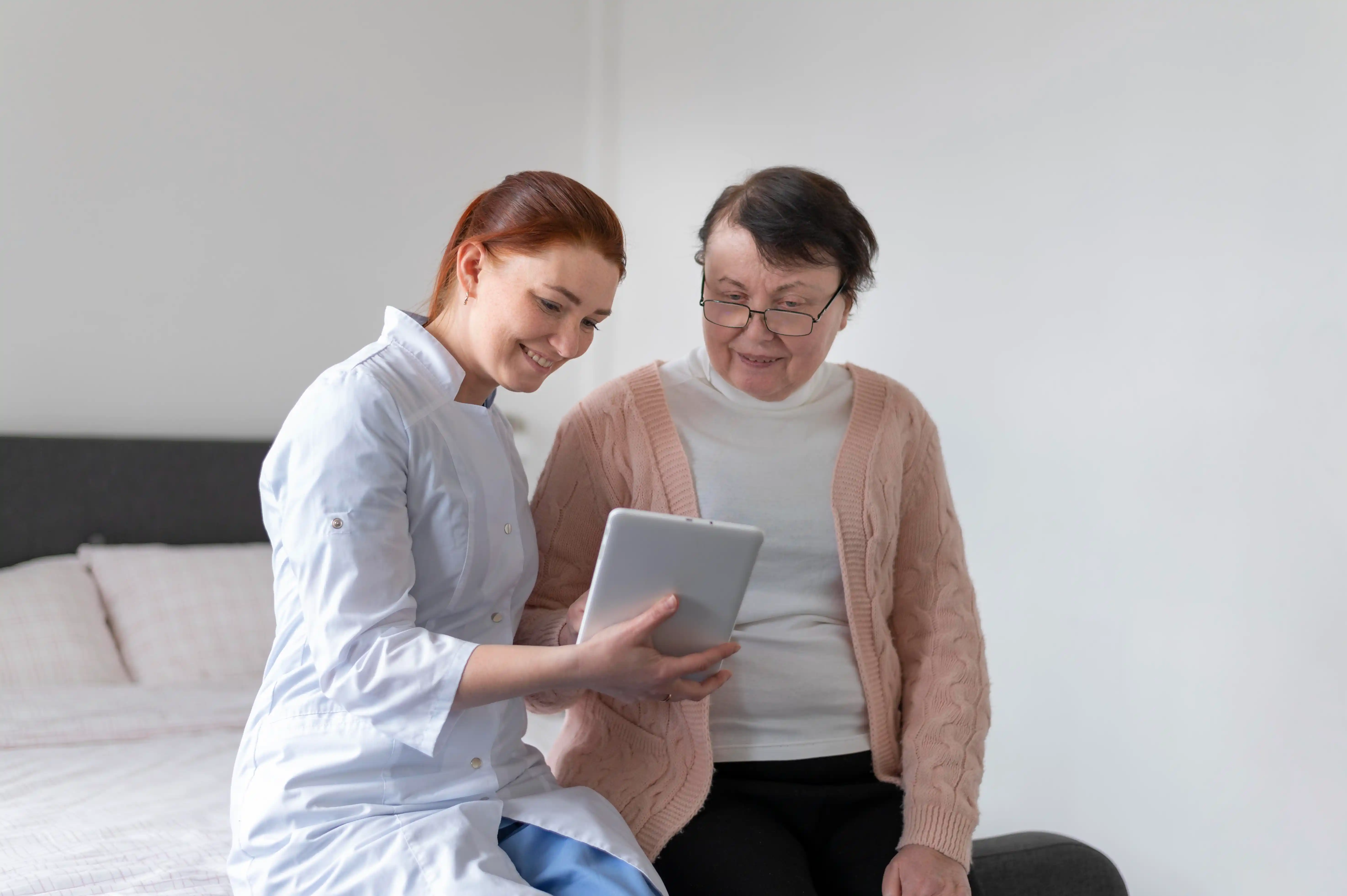A woman in a white coat sits on a bed beside an older woman, both engaged in a conversation.