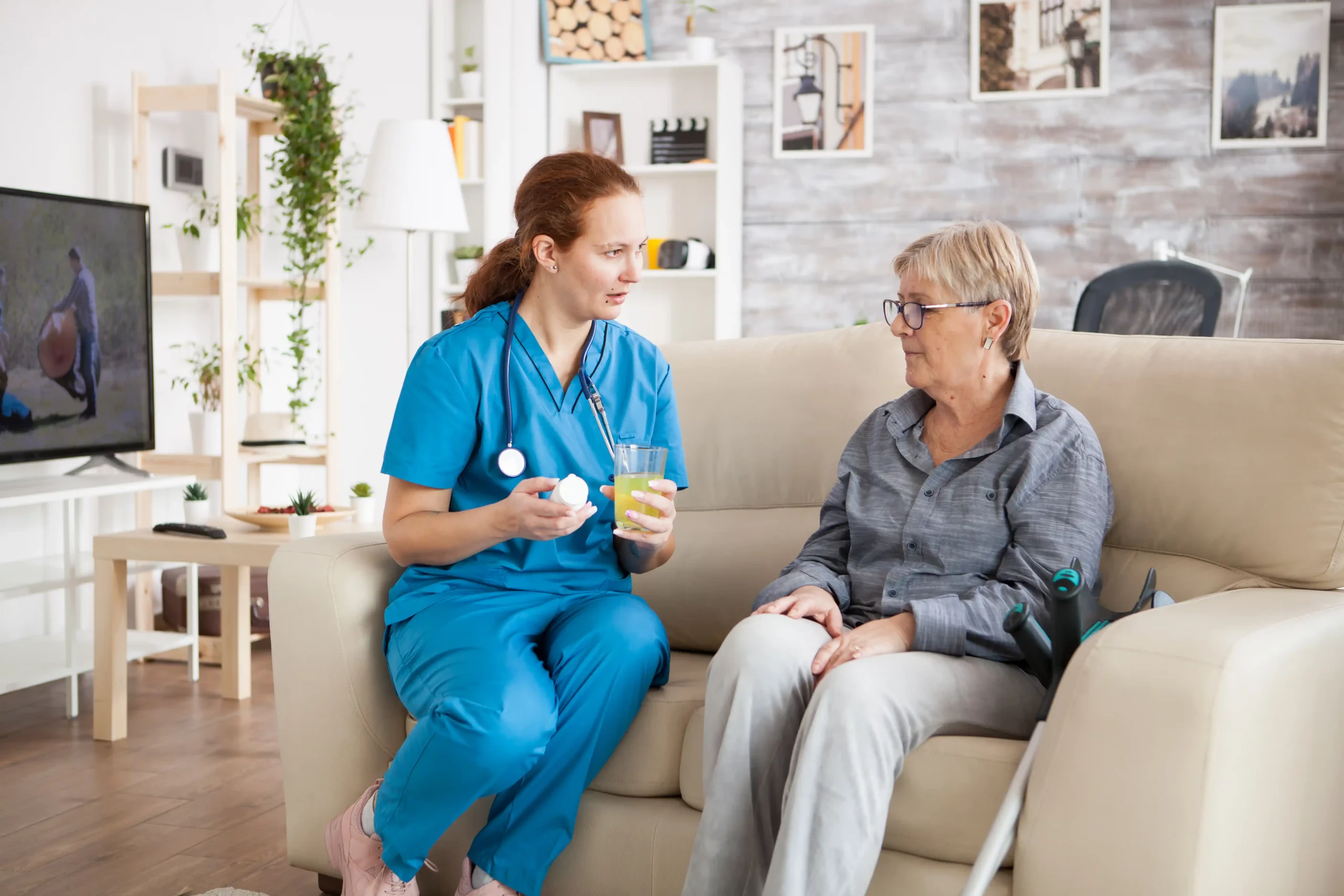 A nurse converses with an elderly woman seated on a couch, providing care and support in a warm environment.