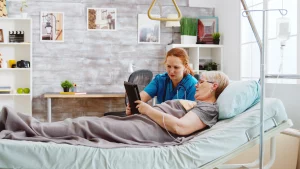 A nurse assists an elderly patient in a hospital bed using a tablet. The room is bright and modern, with a caring atmosphere, books, and plants in the background.