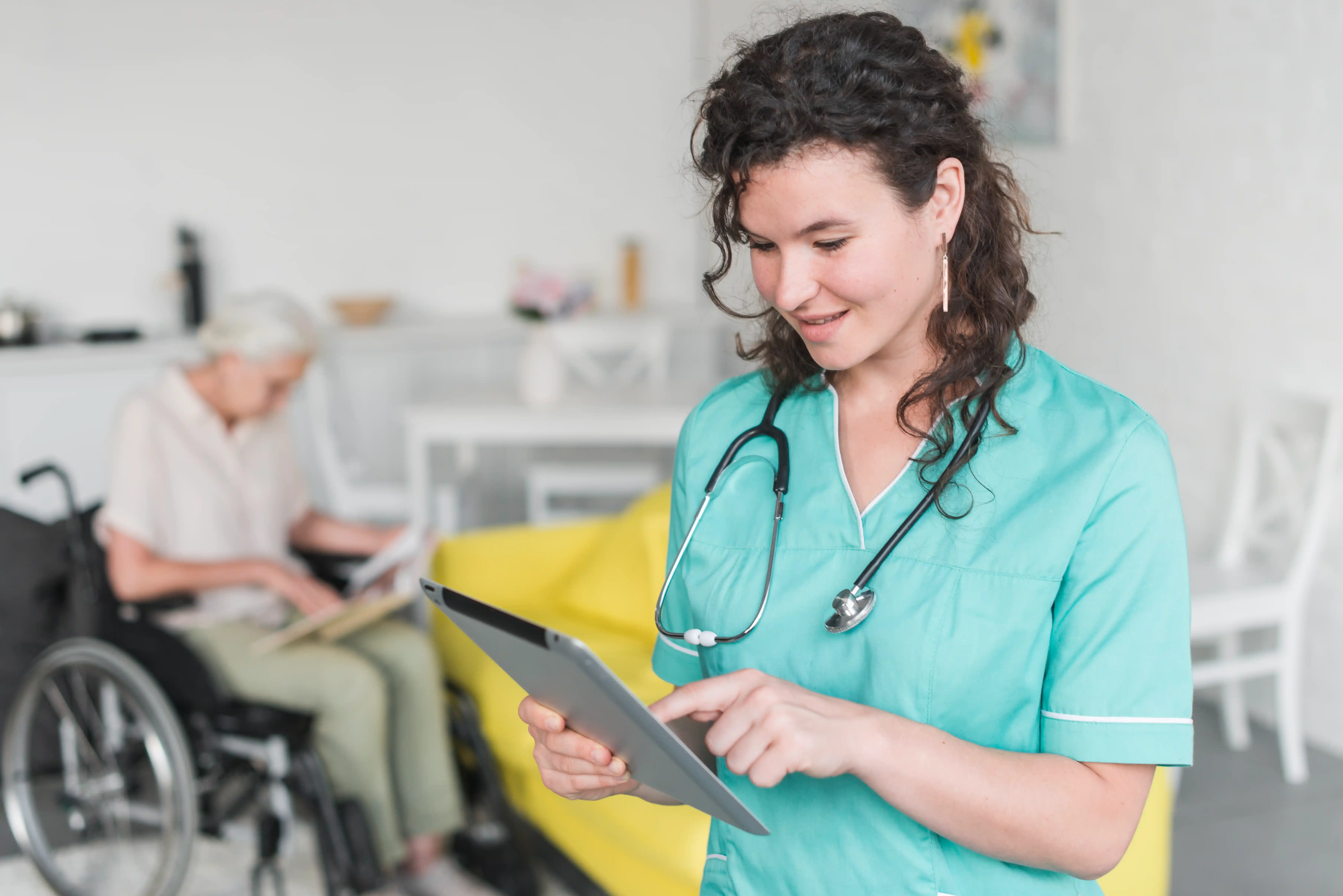 A smiling nurse in a teal uniform uses a tablet, with an elderly person in a wheelchair reading in the background. The setting appears calm and caring.