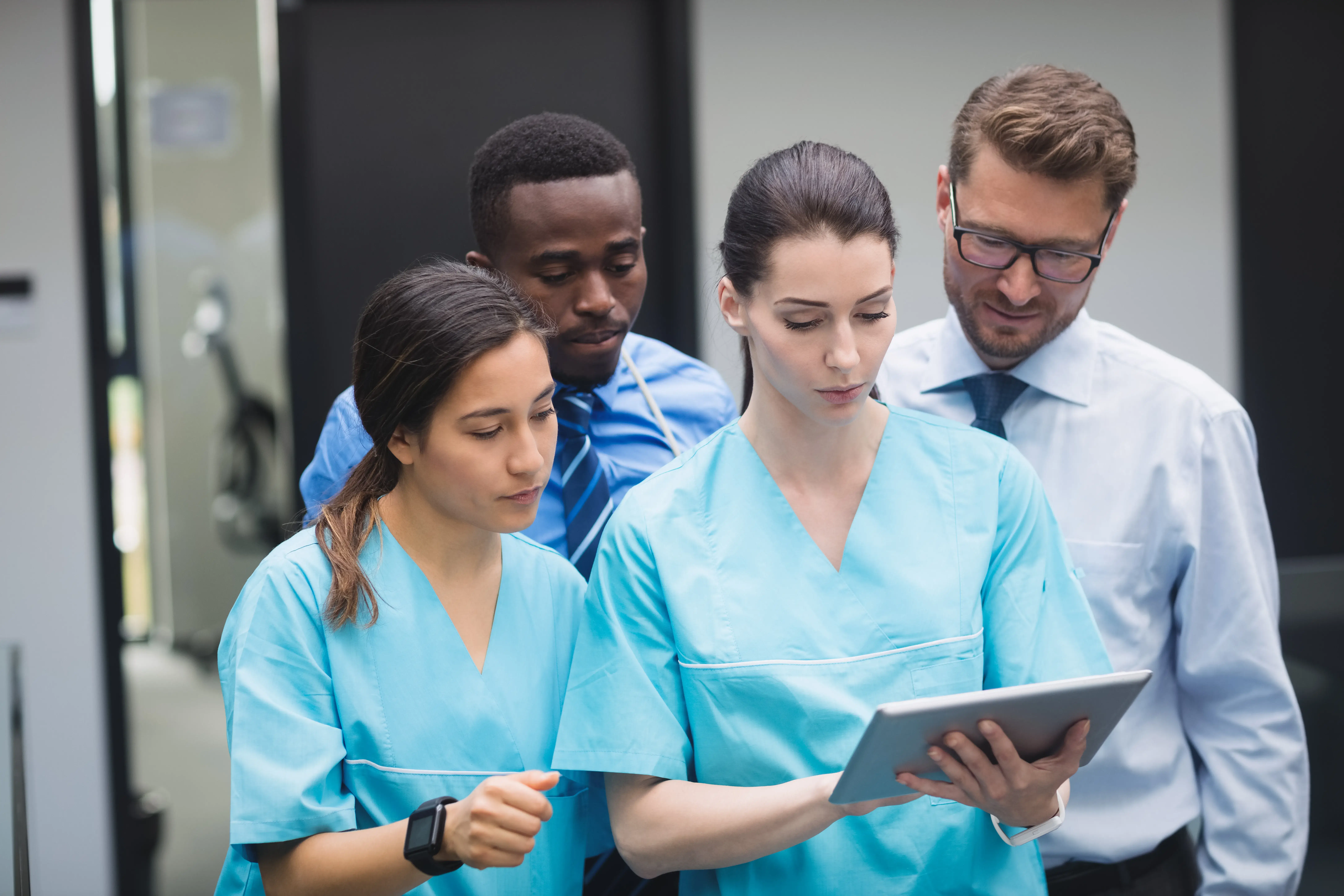 Four medical professionals in light blue scrubs and a man in business attire discuss information on a tablet, conveying focus and collaboration.