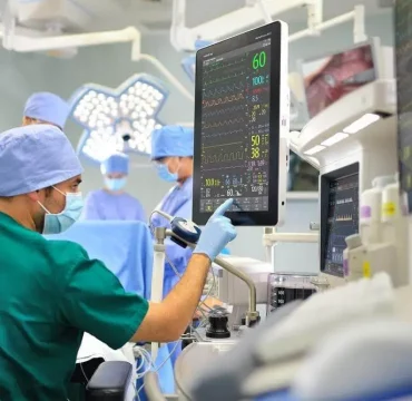 A medical team in blue scrubs conducts surgery in an operating room. A technician in green monitors vital signs on a screen, ensuring patient safety.