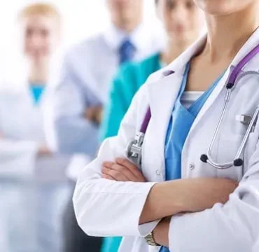A group of healthcare professionals stands confidently; a female doctor in front with folded arms, stethoscope visible, conveying teamwork and professionalism.