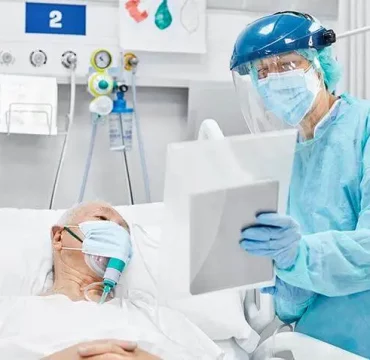 A healthcare worker in full protective gear, including a face shield and mask, checks on an elderly patient with an oxygen mask in a hospital bed.