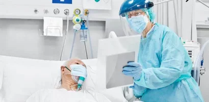 get-yourself-hire A healthcare worker in full protective gear, including a face shield and mask, checks on an elderly patient with an oxygen mask in a hospital bed.